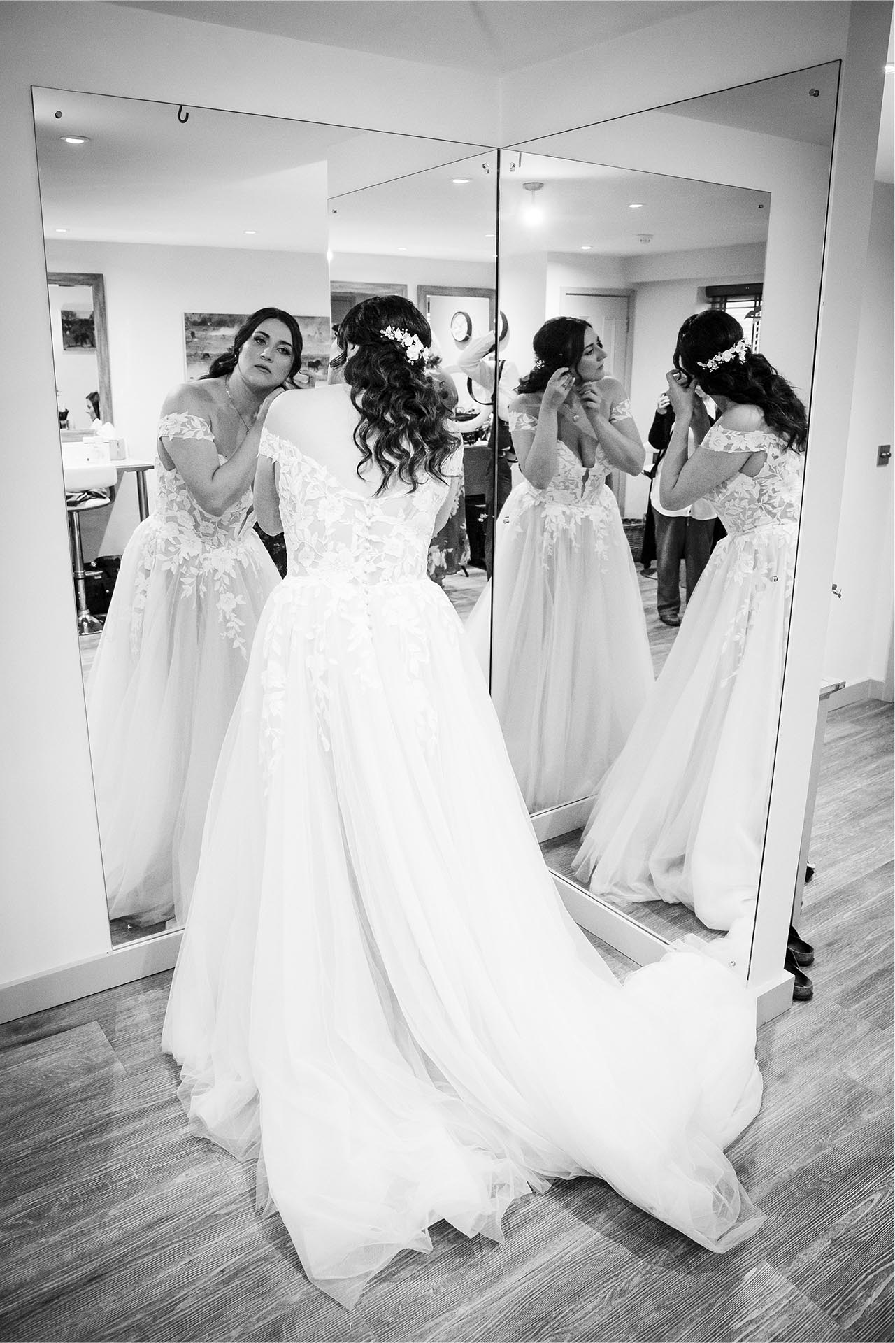 Black and white photograph of bride putting on jewellery in a mirror