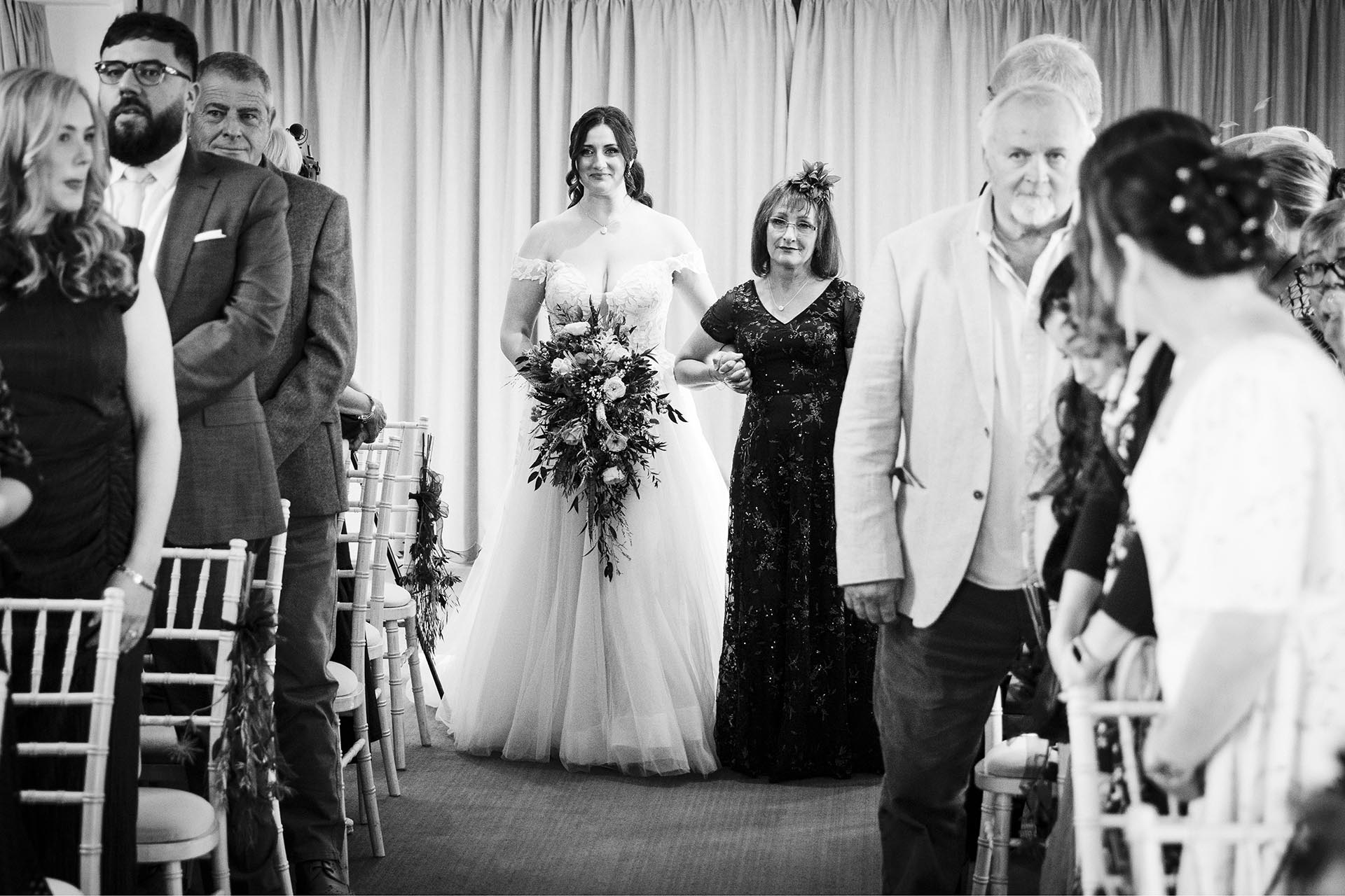 Black and white photograph of bride walking down the aisle with her mother in the ceremony room at The Compasses at Pattiswick, Essex