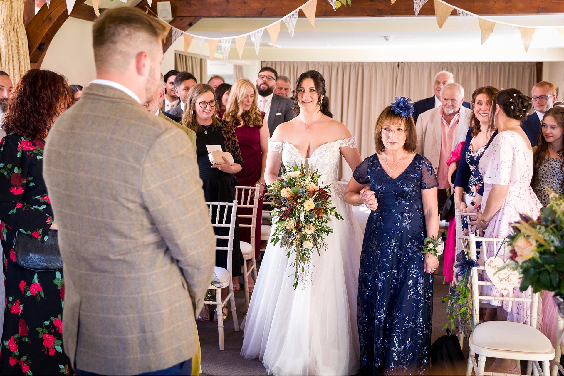 Photograph of bride walking down the aisle with her mother in the ceremony room at The Compasses at Pattiswick, Essex