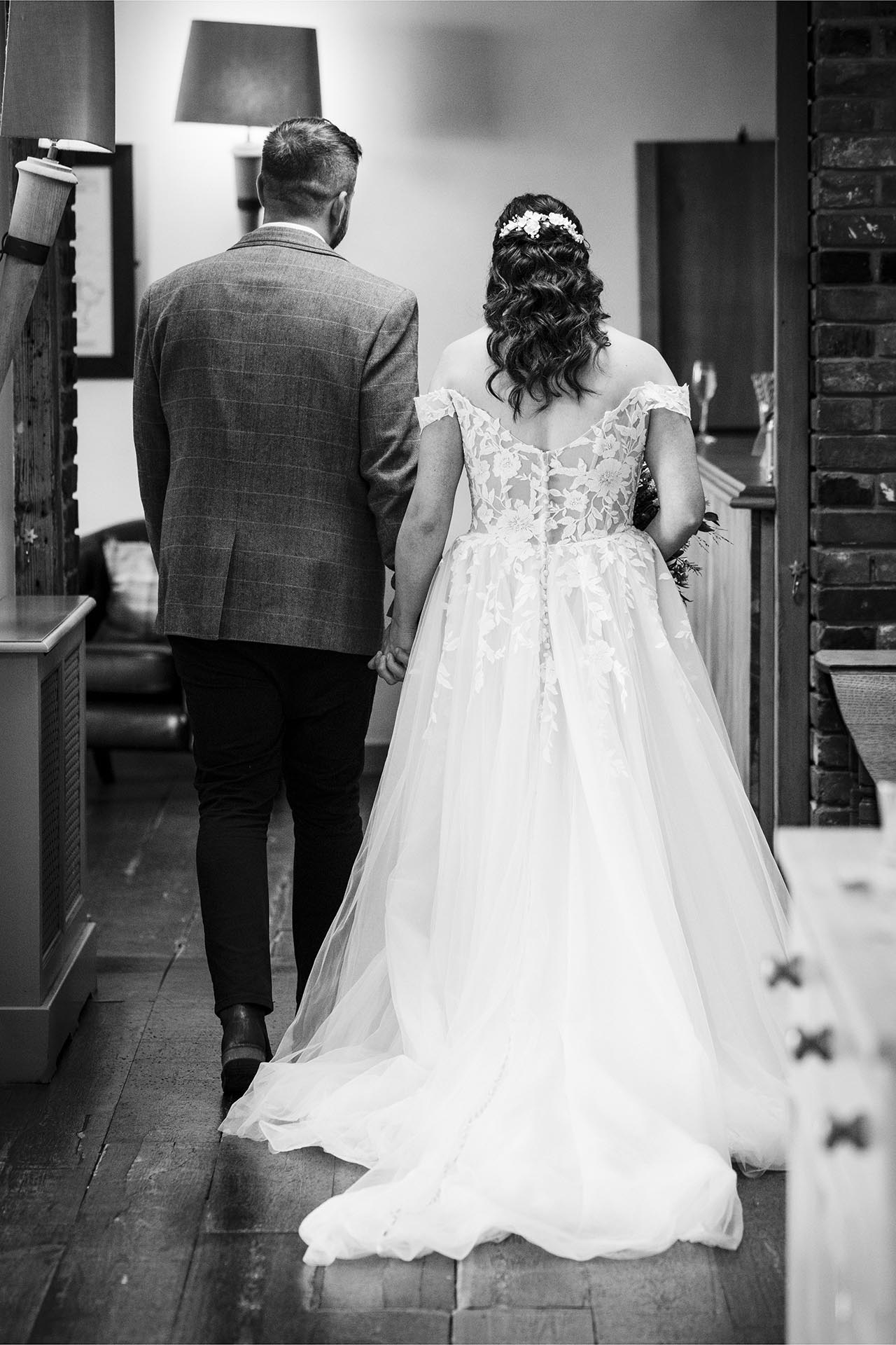 Black and white photograph of the backs of the bride and groom as they leave the ceremony room at The Compasses at Pattiswick, Essex