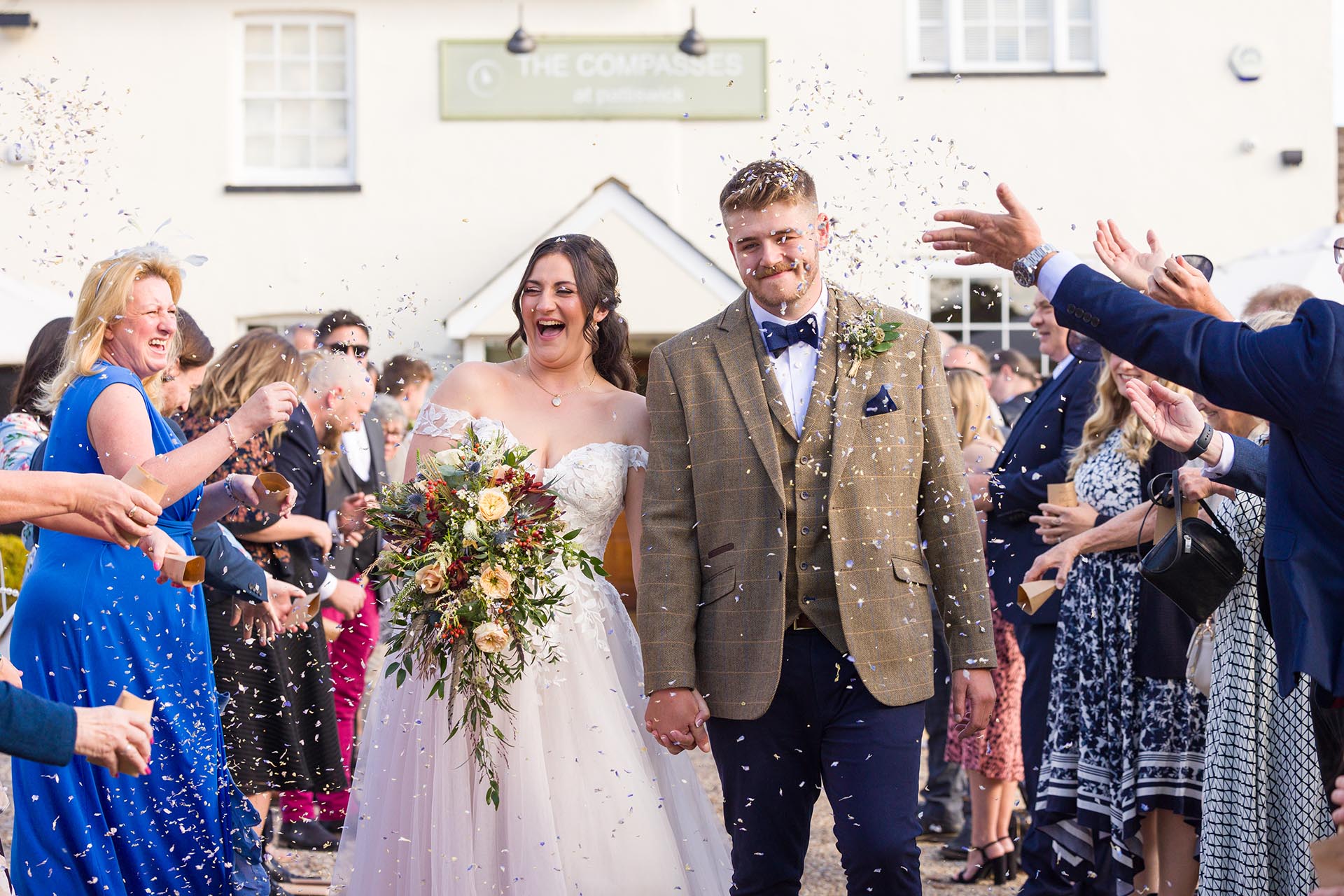Photograph of guests throwing natural flower petal confetti over the bride and groom with The Compasses at Pattiswick wedding venue in the background
