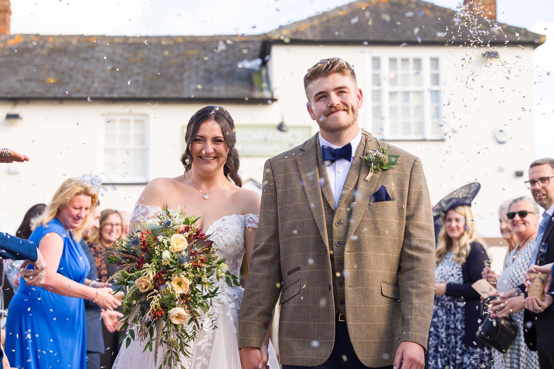 Photograph of guests throwing natural flower petal confetti over the bride and groom with The Compasses at Pattiswick wedding venue in the background