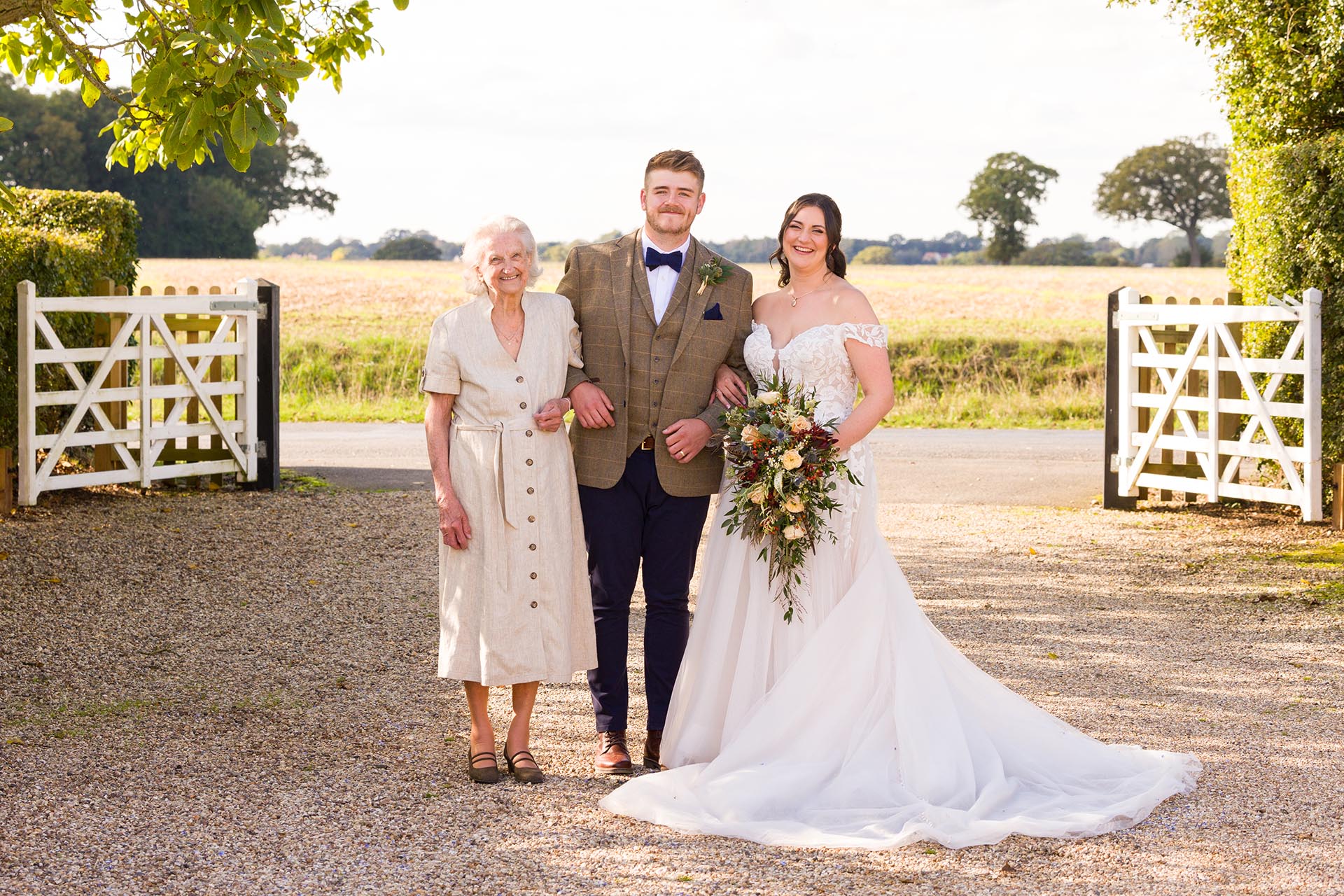 Photograph bride and groom with a guest infront of fields and the white gates at The Compasses at Pattiswick wedding venue