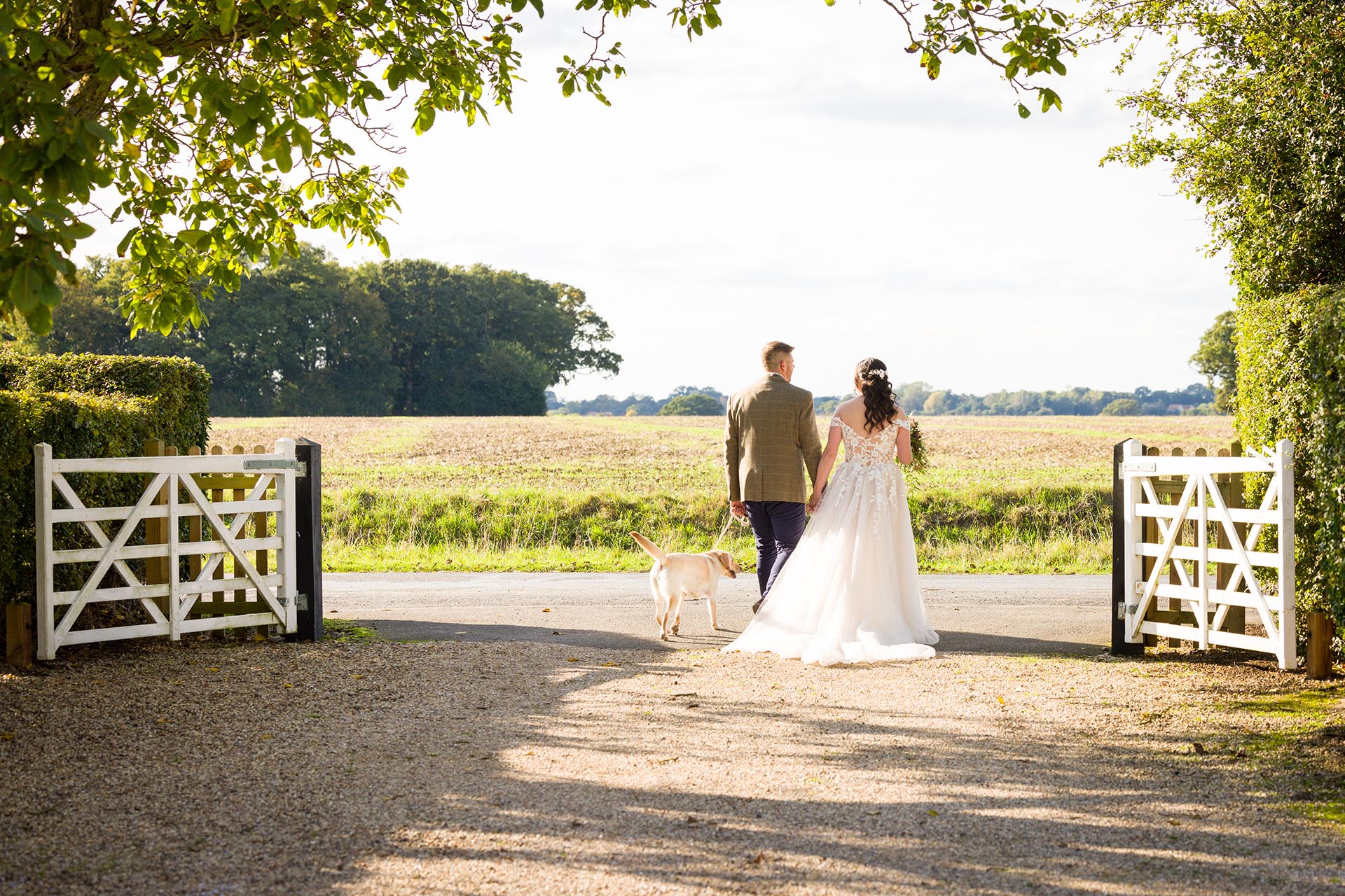 Photograph of bride and groom walking out of the white gates at The Compasses at Pattiswick with their labrador dog