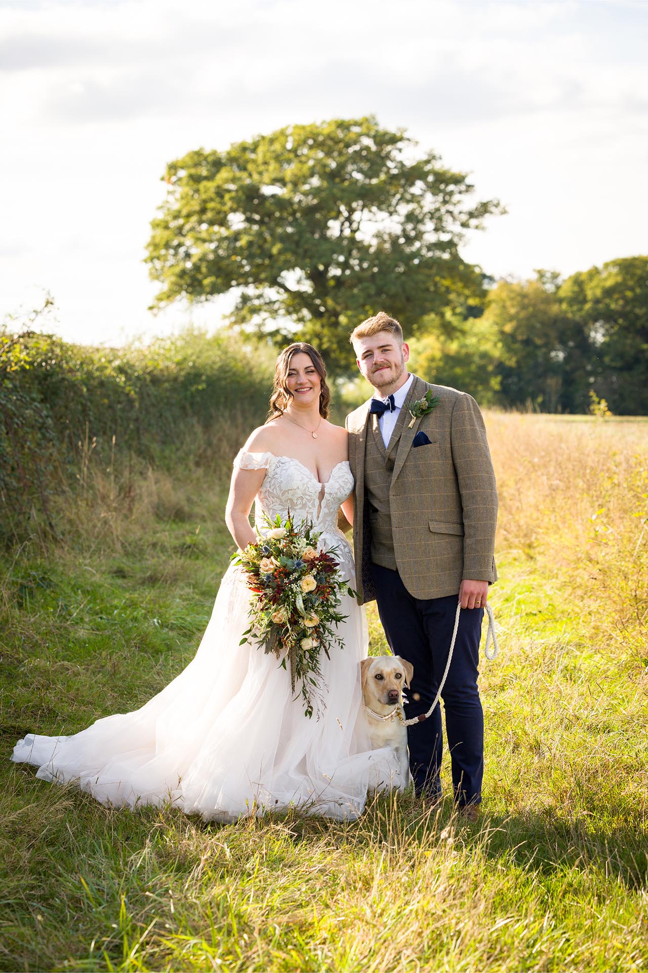 Photograph of bride and groom with their labrador dog in a field near The Compasses at Pattiswick