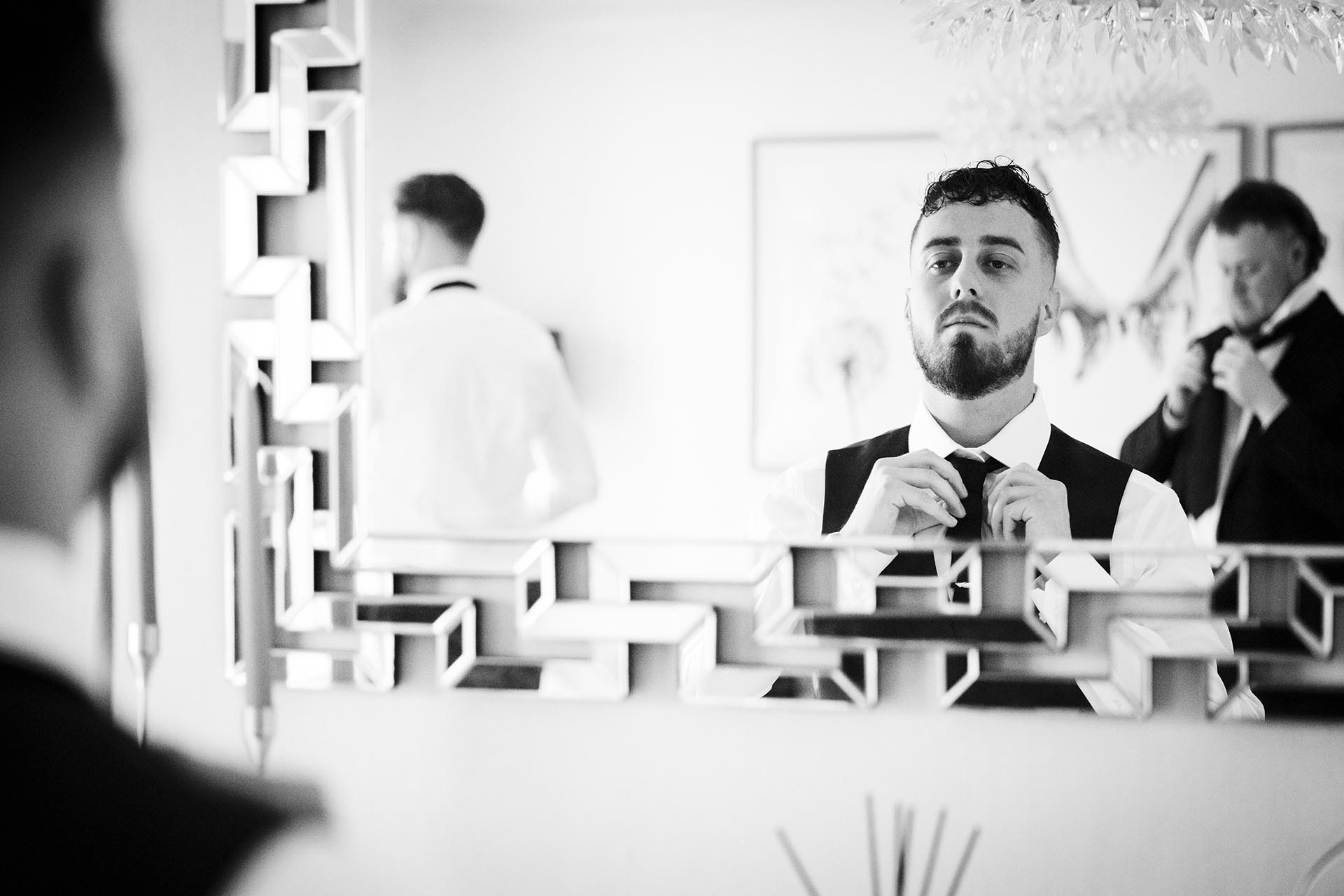 Black and white photograph of groom doing up his tie in a mirror