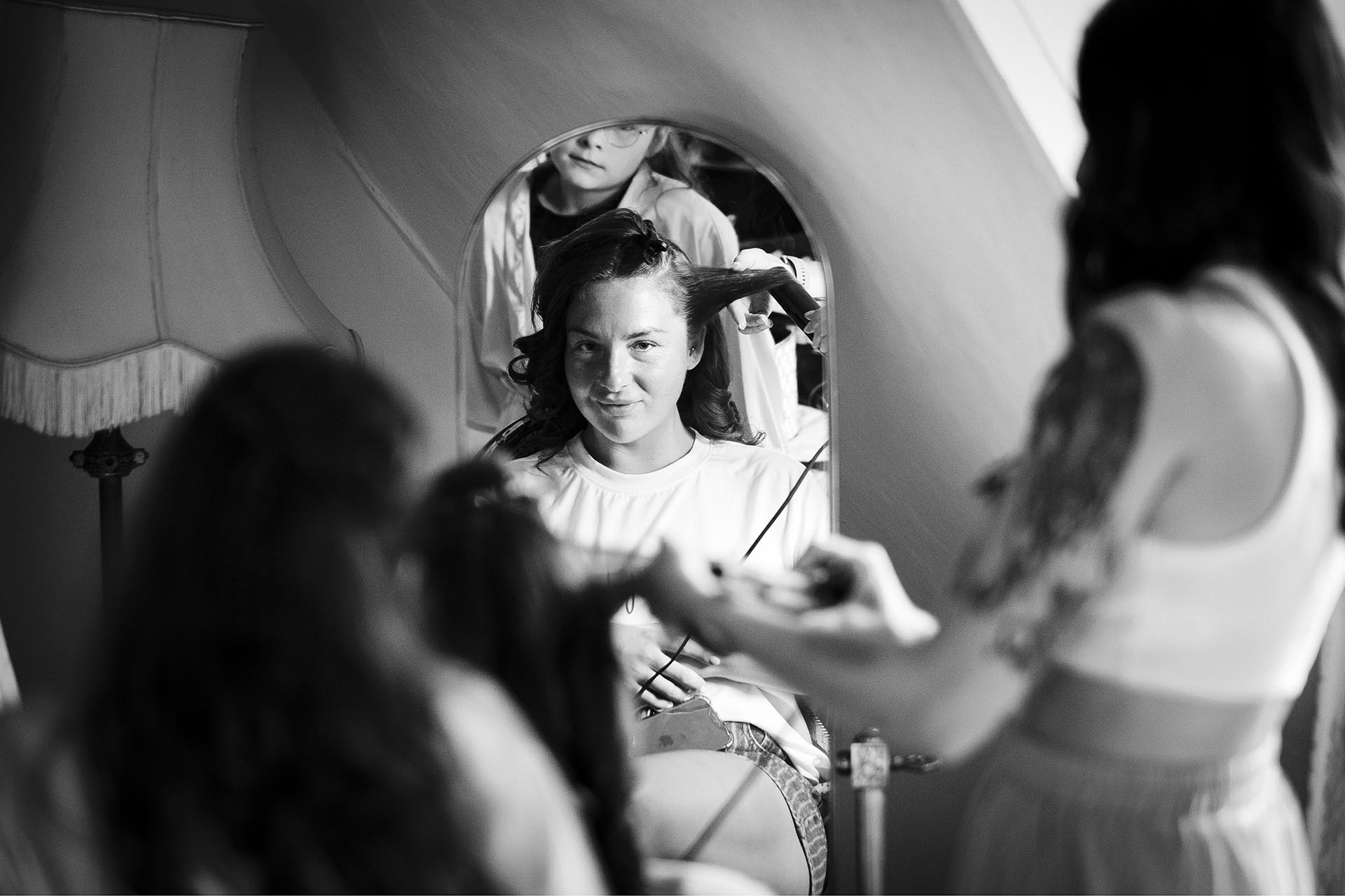 Black and white photograph of bridesmaid looking at herself in a mirror whilst her hair is being brushed