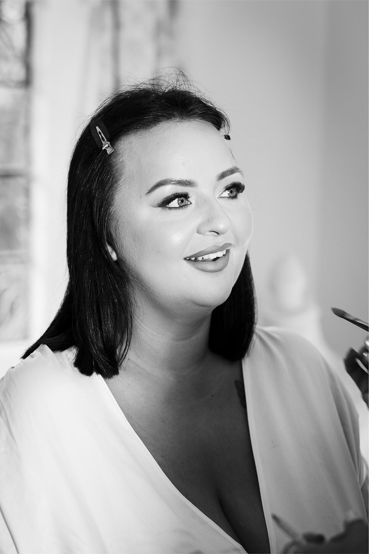 Black and white photograph of bride smiling whilst having make-up applied