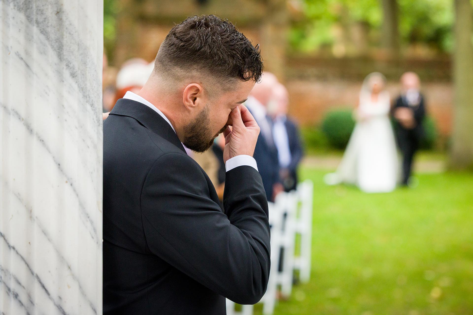 Photograph of groom wiping tears from his eyes as bride walks down the aisle with her father for outdoor wedding ceremony