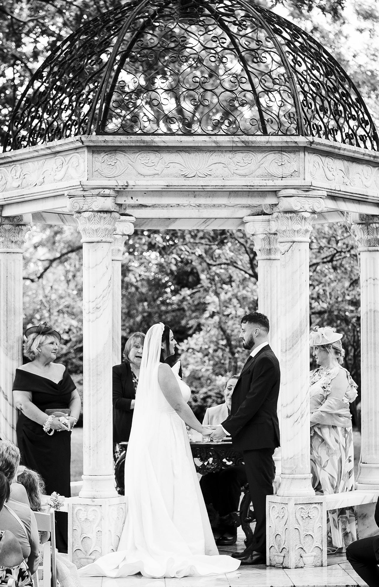 Black and white photograph of bride and groom holding hands and looking at each other under the Marble Gazebo during outdoor wedding ceremony in the orchard at Creeksea Place, Essex