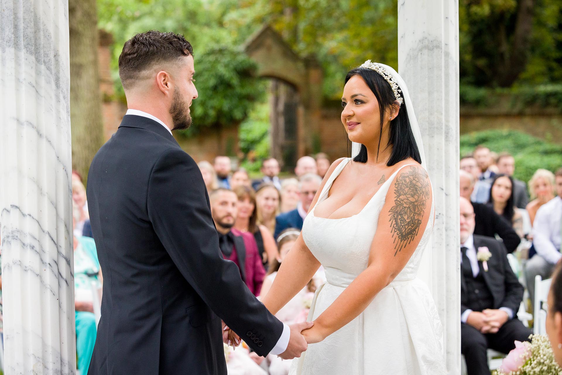 Photograph of bride and groom holding hands and looking at each other during outdoor wedding ceremony at Creeksea Place, Essex
