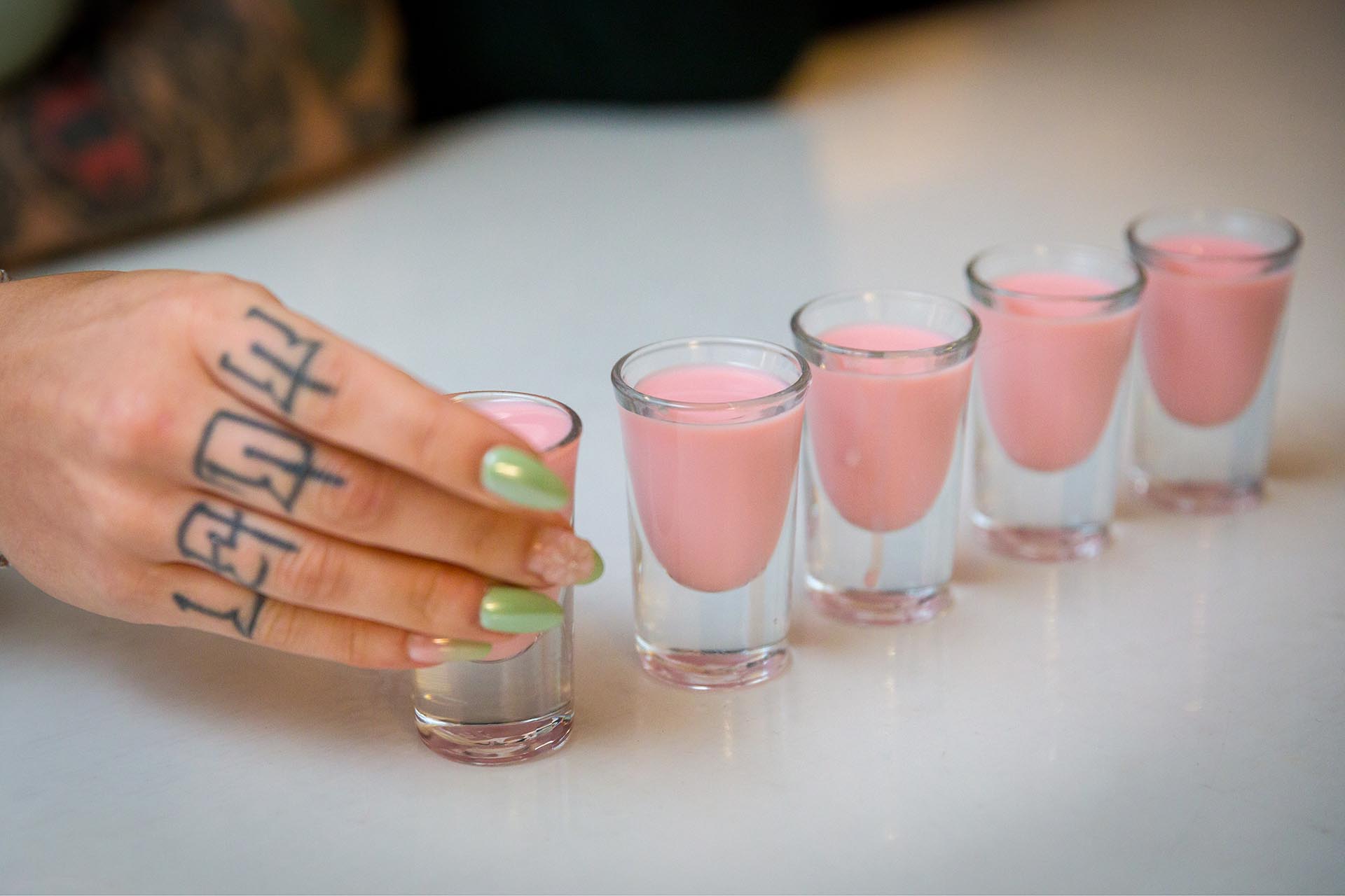 Photograph of a hand picking up one of a line of shot glasses containing pink drink
