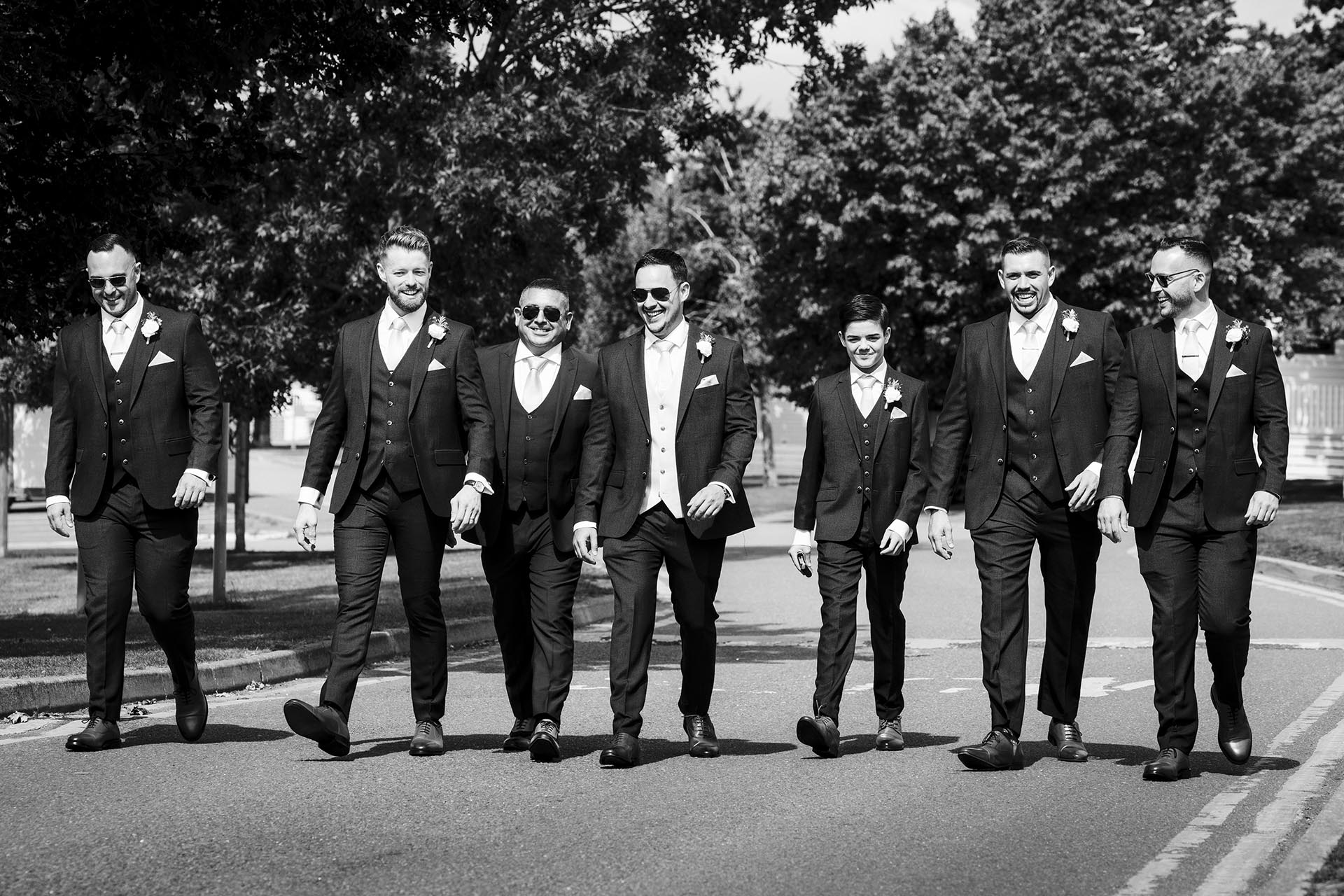 Black and white photograph of groom and groomsmen walking along a road laughing