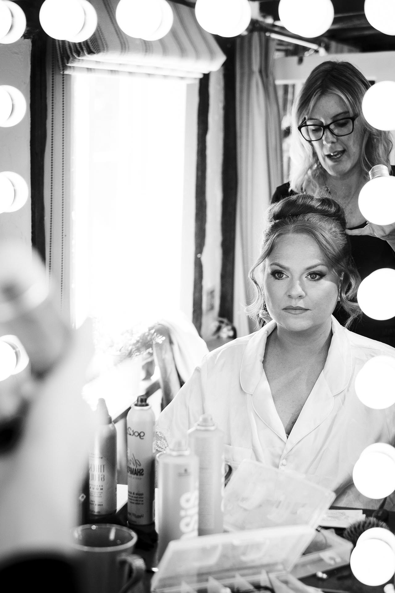 Black and white photograph of the bride looking at herself in a mirror whilst her hair is being done