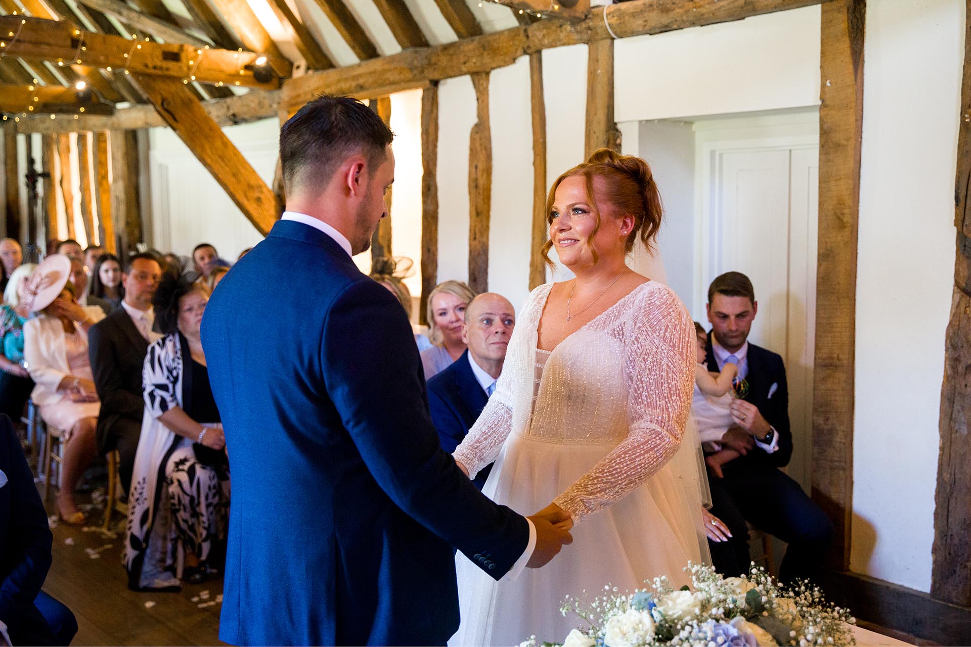 Bride and groom smiling at each other whilst holding hands in front of a full ceremony room at Little Channels