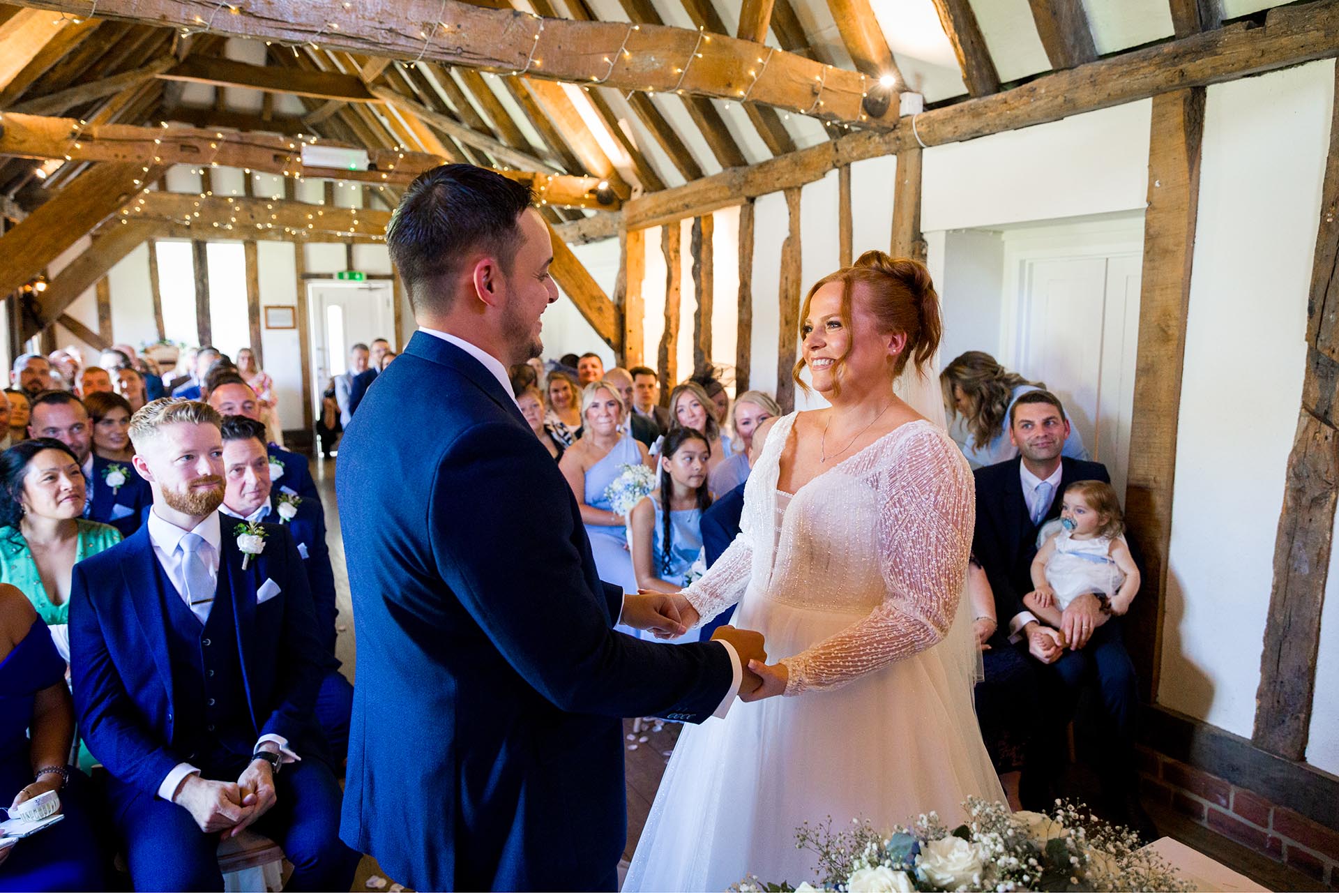 Bride and groom smiling at each other whilst holding hands in front of a full ceremony room at Little Channels