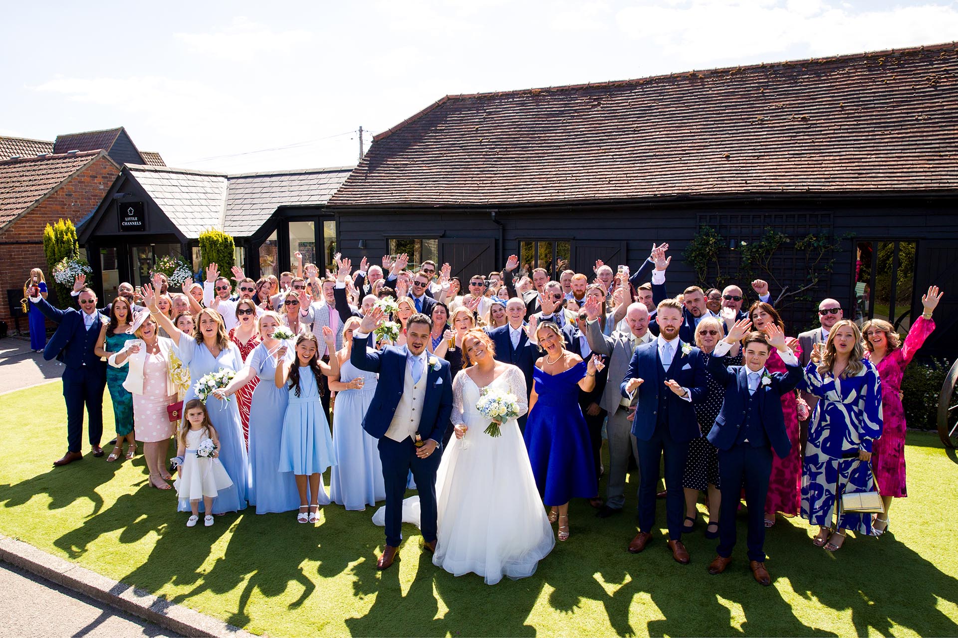Group photograph of bride and groom waving at the camera with all of their guests
