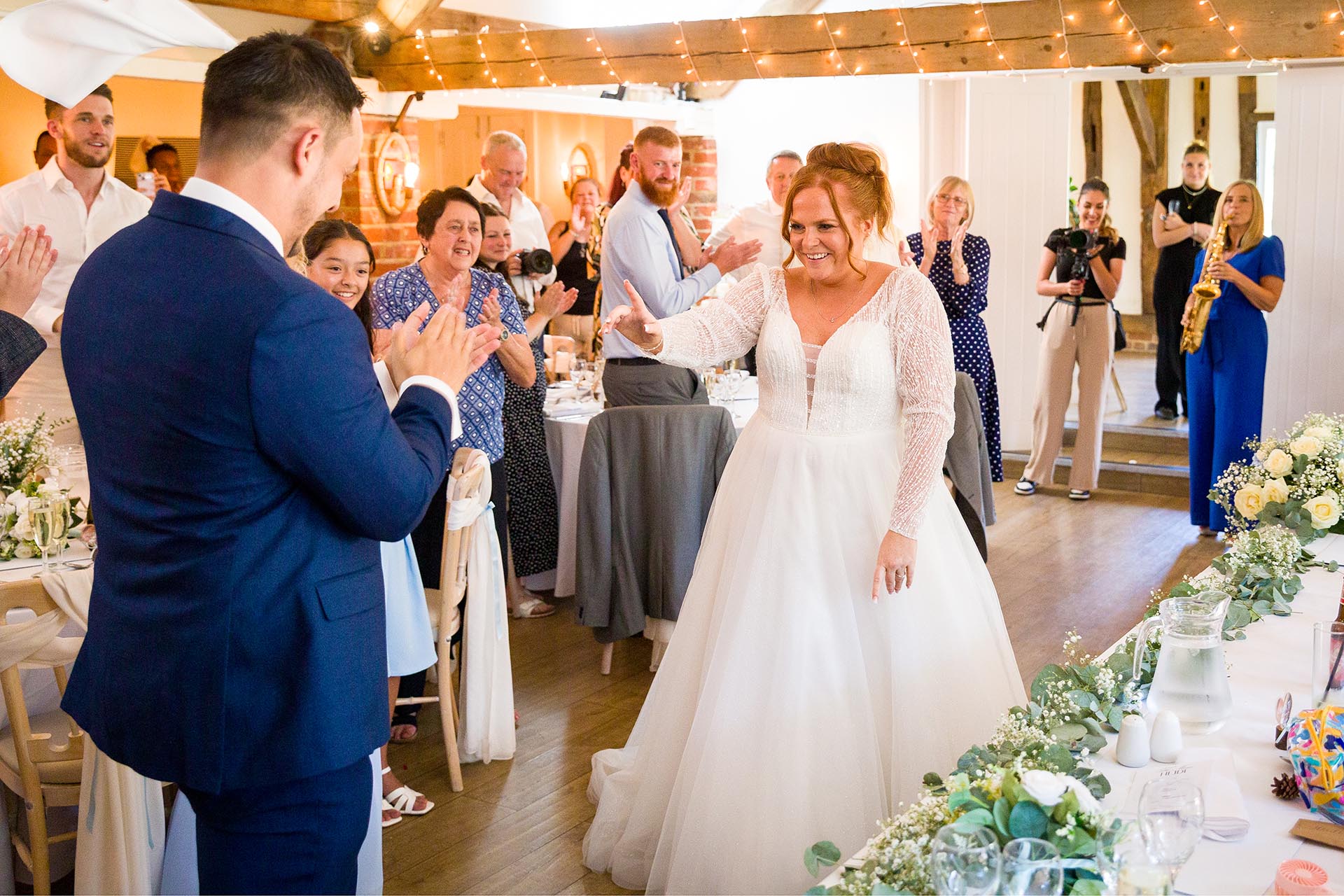 Bride and groom laughing and dancing as they enter their wedding breakfast room with guests applauding and a saxophonist playing in the background