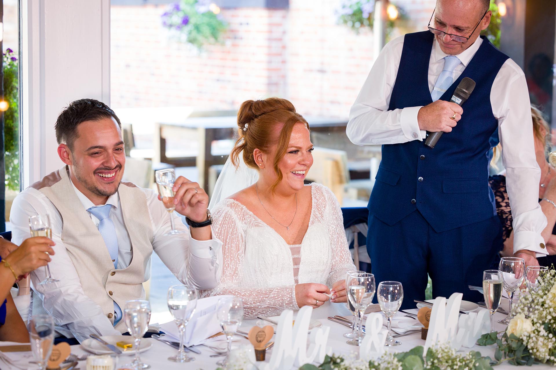 Bride and groom laughing as bride's father looks on whilst making his speech