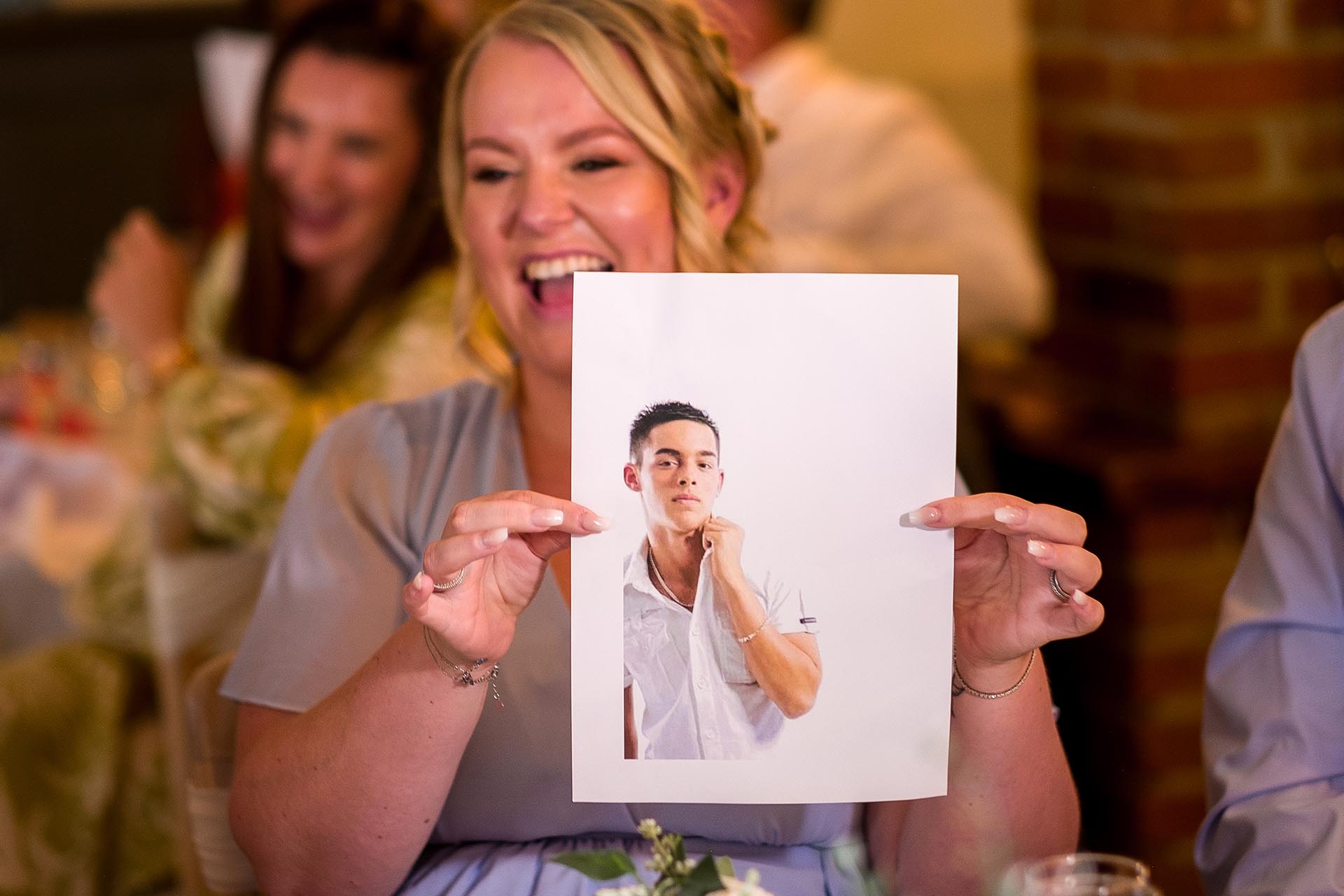A laughing guest holding up a photograph of the groom when he was younger