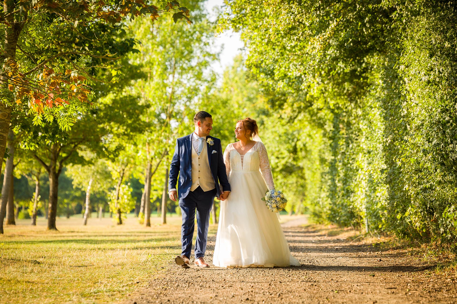 Bride and groom holding hand and looking at each other whilst walking along a tree-lined path at Little Channels, Essex