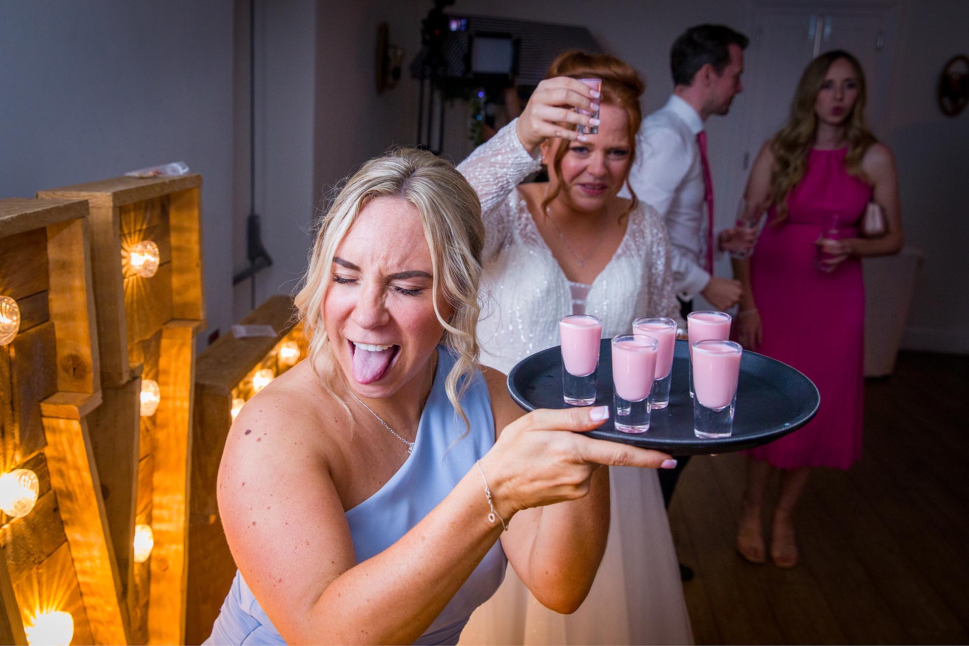 A bridesmaid pulling a face with her tongue out as the bride raises a shot glass