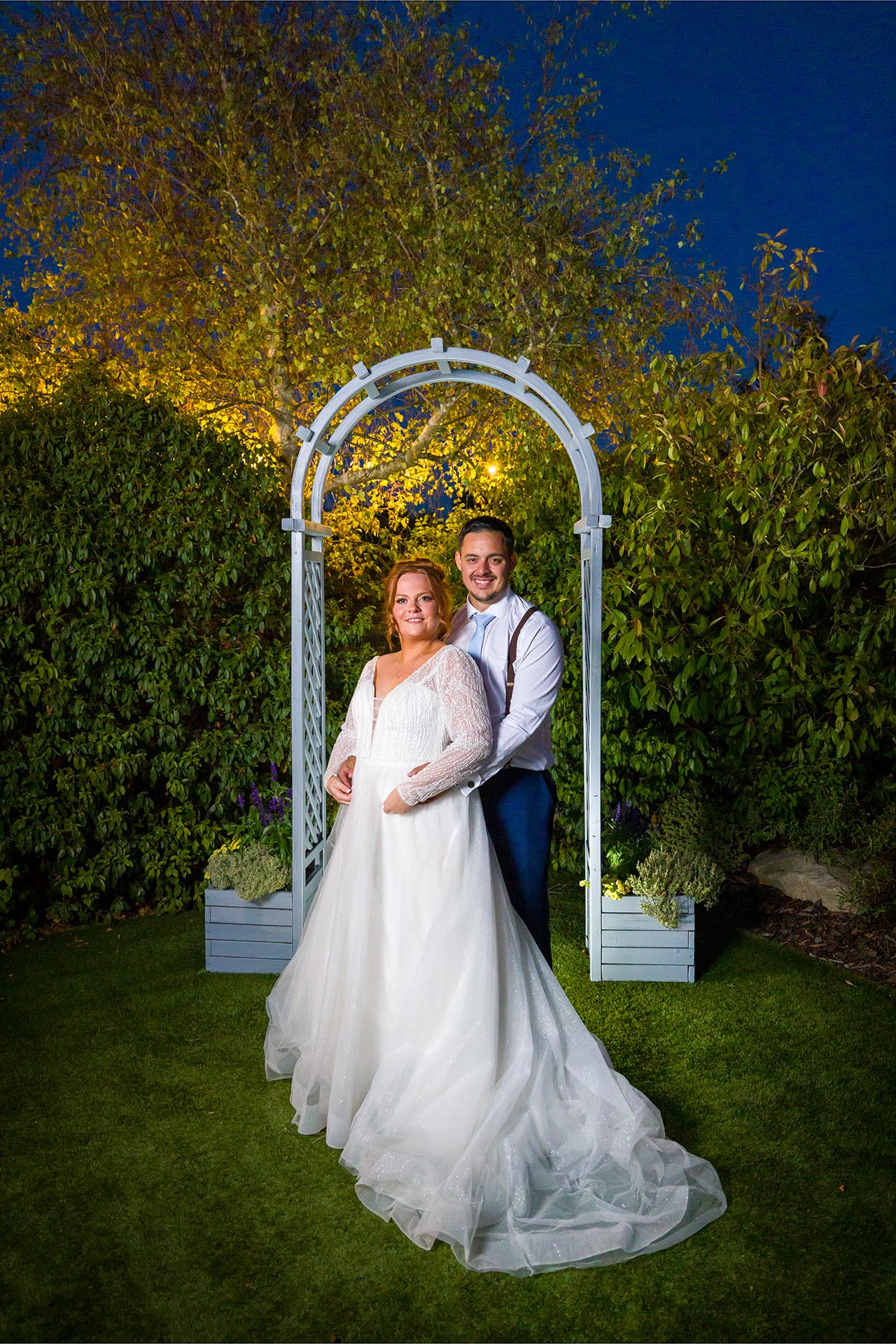 The bride and groom smiling at the camera at twilight under a garden arch at Little Channels, Essex